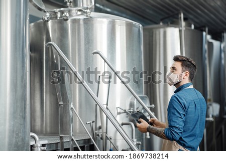 Modern brewery, craft beer and covid-19 quarantine. Young male worker in apron and protective mask with tablet in hands works with equipment in interior of plant with kettles, profile, free space