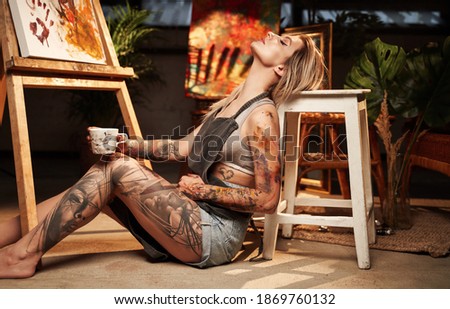 Attractive tattooed female designer with blond hairs and apron poses on floor holding coffee cup in dark atmospheric workshop.