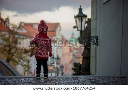 Beautiful toddler child with lantern and teddy bear, casually dressed, looking at night view of Prague city, wintertime