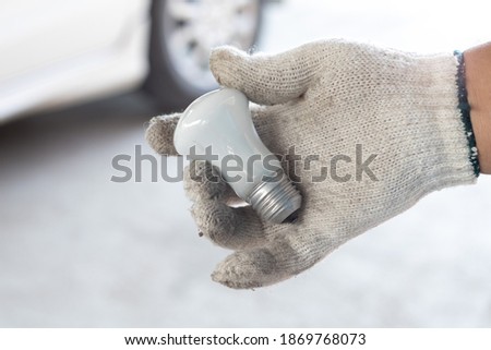 Hand wearing white gloves  Holding a light bulb, A white car in the background