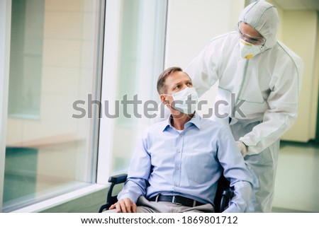 Ill senior man with protective safety mask on face in a wheelchair and a confident doctor in the medical mask while transporting on the hospital.