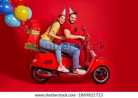 funny two young people friends drive motorbike deliver gifts presents for birthday, isolated red color studio background, happy cheerful man and woman having fun carrying festal balls