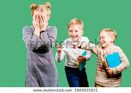 Children two boys bullying poor girl, sneering, offending her at classroom. The poor kid is covering face with hands sobbing. Concept of aggression towards other child. Royalty-Free Stock Photo #1869835831