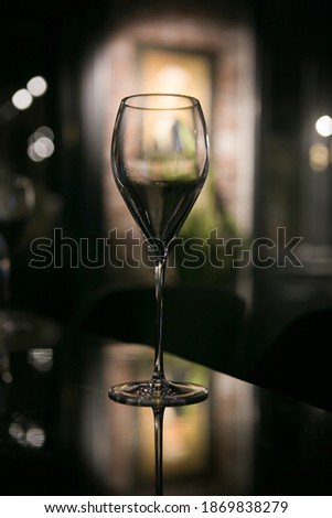A glass of champagne on a mirror surface. In dark colors with reflection. In the background there is a picture and greenery.

