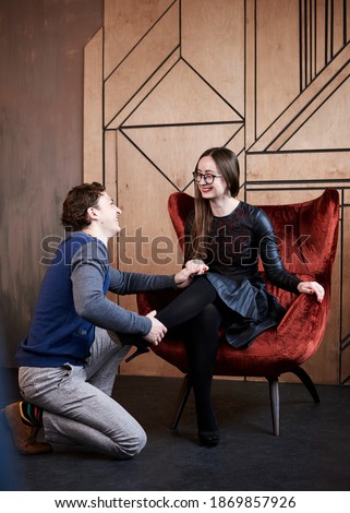 Young woman, sitting on red velvet Santa armchair and man, kneeling down near her in front of wooden wall, smiling, laughing. Romantic relationship. Young couple in fancy clothes, posing for picture.