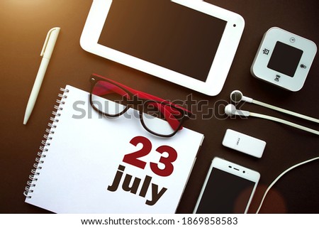 July 23rd. Day 23 of month, Calendar date. Office workplace with laptop, notebook, office supplies and stationery on brown back. Summer month, day of the year concept