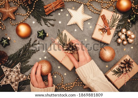 Christmas and New Year rustic traditional card. Festively decorated cedar branches on a gray concrete board. Craft handmade gift boxes, golden balls, stars and cones. Female hands wrapping a gift.