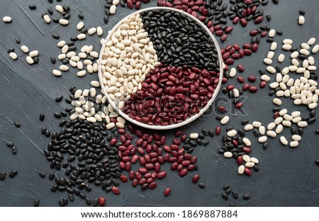 beans in white ceramic plate. bowl of beans. black bean, red kidney bean and white bean. navy, cannellini, white kidney bean on dark gray background and scattered various beans