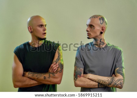 Horizontal shot of two young caucasian twin brothers with tattoos and piercings keeping arms crossed and looking at each other while posing together in studio. Family and friendship concept