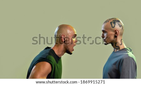 Portrait of two young men, twin brothers with tattoos and piercings arguing, shouting, standing face to face isolated over light green background. Side view. Web Banner