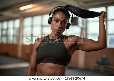 Ethnic female athlete looking away and pulling ponytail while listening to music during break in training in modern gym