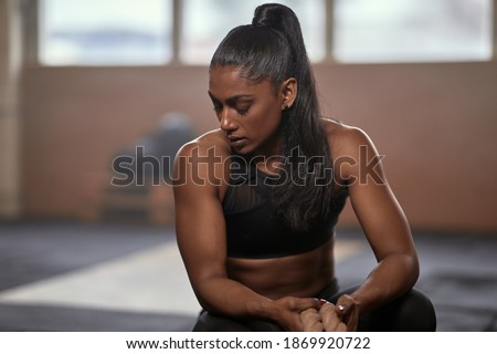 Exhausted ethnic female athlete with rope sitting on haunches and breathing heavily with closed eyes during break in training on blurred background of gym Royalty-Free Stock Photo #1869920722