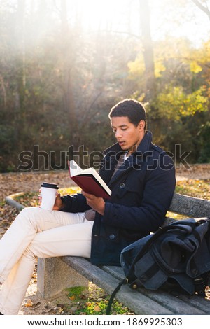 Stock photo of a young man sitting on a bench reading a book and drinking coffee outdoors in nature.
