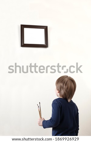 Handsome caucasian boy with thick hair hold the paintbrushes standing in front of white free space in horizontal frame on the wall and looks up to it, close up