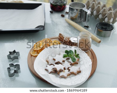 Ingredients and kitchen utensils on the kitchen glass table. Cookies on a white plate on a wooden tray. Christmas cookies decorated with powdered sugar and a sprig of mint