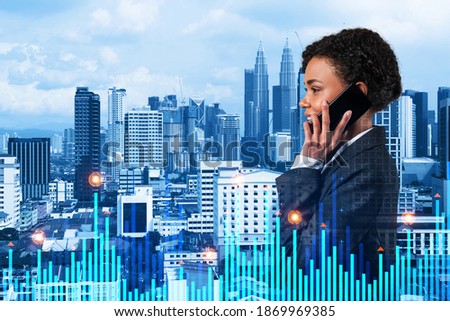 Attractive black businesswoman pensively processing conference call by phone to find financial solution for venture capital project. Hologram chart over Kuala Lumpur city background