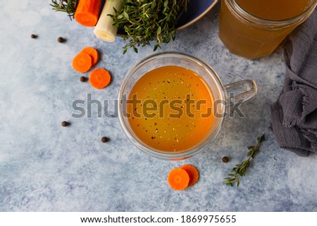 Homemade bone broth in glass mug and vegetables, blue concrete background. Collagen source for the body. Selective focus. Top view.