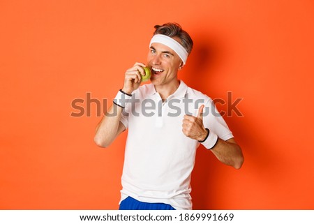 Portrait of healthy and active middle-aged man, eating an apple and showing thumbs-up, standing against orange background