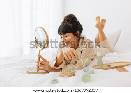 Close up of smiling young woman lying on bed and applying face cream. Woman doing beauty care face massage at home. Royalty-Free Stock Photo #1869992359