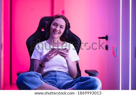 Gamer girl in a big professional music headphones in white t-shirt and blue jeans holds smart phone in hands and plays game app on mobile phone, smiling and looking at camera indoors.