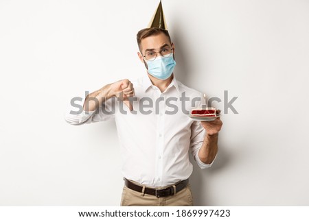 Coronavirus, quarantine and holidays. Man showing thumb down as disappointed with birthday party, wearing face mask and holding bday cake, white background