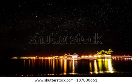 Night on tropical resort, many little beach houses glowing bright yellow lights in dark night, starry sky over sea, romantic summer holidays concept