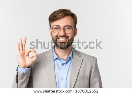 Close-up of handsome confident businessman, wearing glasses and gray shirt, showing okay sign and smiling, approve something, standing over white background