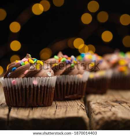 Tasty chocolate cupcakes with sprinkling on rustic wooden table on black background with yellow lights. Sweet dessert. Bakery concept. Elegant food. Sweets for coffee or tea. Space for text.