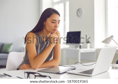 Artist looking at laptop screen and thinking while drawing on graphic tablet. Pensive web designer working on project. Young woman editing and retouching images using digital tablet and stylus pen