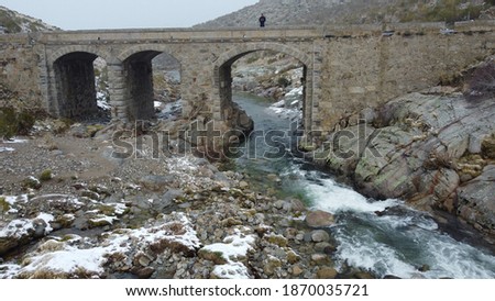 
photo of a snowy bridge with snowy ice with a person and a river below the bridge with a forest in the background
