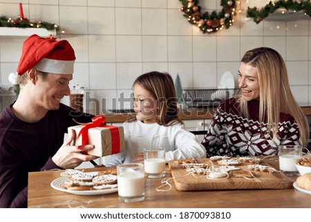 Happy dad wearing santa hat giving excited cute small kid daughter xmas gift sitting at kitchen table. Young parents preparing present for child girl celebrating Christmas family holiday at home.