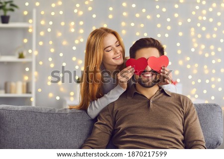 Happy smiling couple in love celebrating their relationship anniversary at home. Young people having fun on Saint Valentine's Day. Woman covering boyfriend's eyes with two red heart-shaped cards Royalty-Free Stock Photo #1870217599