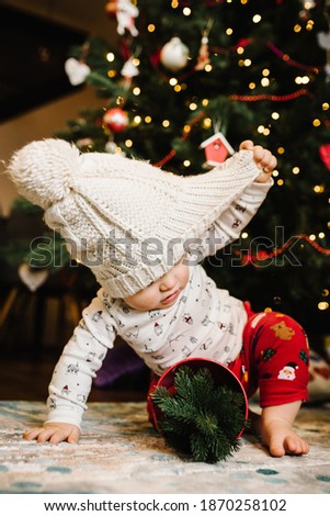 Toddler boy in a winter hat and Christmas costume sitting on the floor in room. The child plays with toys on the background of the Christmas tree. Happy holidays. Happy New Year and Merry Christmas.