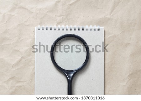 Gray notepad with white coiled spring and magnifier on a background of beige crumpled craft paper. With empty space for text and design