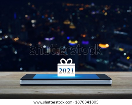 Gift box happy new year 2021 flat icon on modern smart mobile phone screen on wooden table over blur colorful night light city tower and skyscraper, Business happy new year 2021 shop online concept