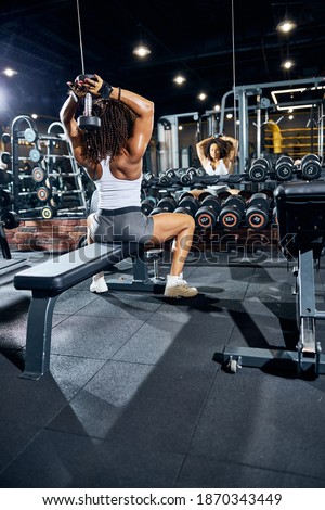 Back view of a muscular curly-haired woman doing the dumbbell overhead triceps before the mirror