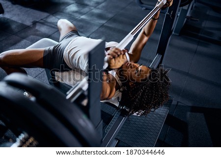 Beautiful serious focused curly-haired young female bodybuilder doing the bench press exercise at the gym Royalty-Free Stock Photo #1870344646