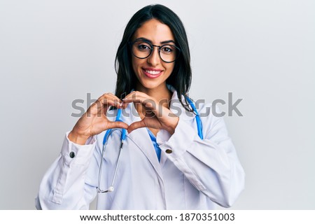 Beautiful hispanic woman wearing doctor uniform and stethoscope smiling in love doing heart symbol shape with hands. romantic concept. 