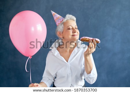 Studio image of fashionable elegant mature lady with short gray hair enjoying birthday party, posing isolated with pink air balloon, closing eyes, eating sweet eclair. Holidays and celebration