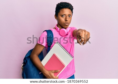 Young african american girl wearing school bag and holding books pointing with finger to the camera and to you, confident gesture looking serious 