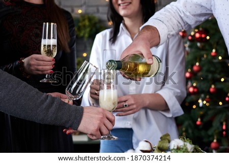Close-up picture of hands with glasses, and bottle of champagne, Christmas tree on background. New year party celebration with friends and family at home.