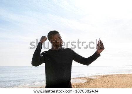Young black sportsman on the beach who shows his strength while making a video call with his cell phone.