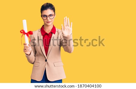 Young beautiful woman wearing glasses holding graduate degree diploma with open hand doing stop sign with serious and confident expression, defense gesture 