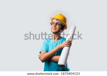 Cute Asian Indian happy kid wearing yellow construction helmet or safety hard hat, standing isolated on white background holding blueprint Royalty-Free Stock Photo #1870404523