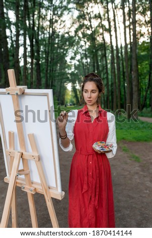 Girl painter in forest. A woman in a red dress, draws a picture, in summer on nature in park, trees background. Easel picture paint and paintbrush. Inspiration and creativity.