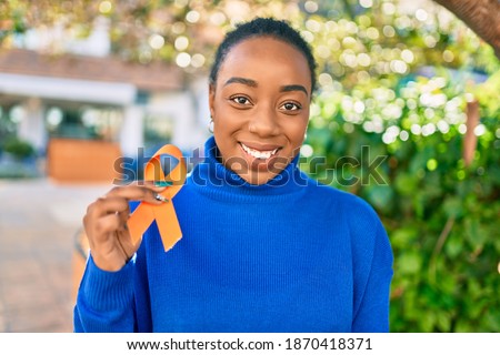 Young african american woman smiling happy holding orange awareness ribbon at the park. Royalty-Free Stock Photo #1870418371