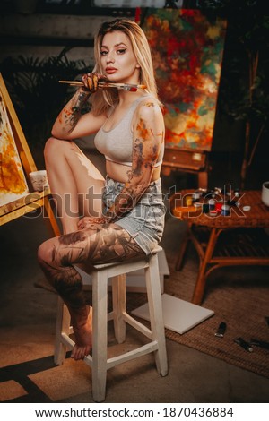 Stylish and beautiful female painter in casual clothing holding paintbrushes and sitting on chair in dark room with canvas and artwork.
