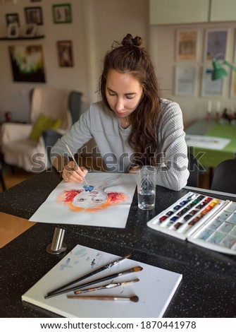 Woman at home at table, draws paints, portrait of a girl. Bright orange paints, color palette. Black table, brush painting. Home studio creativity and art concept.