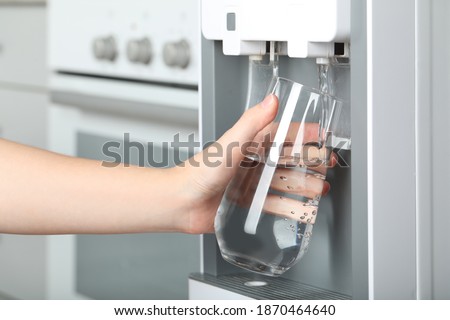 Woman filling glass with water cooler indoors, closeup. Refreshing drink Royalty-Free Stock Photo #1870464640