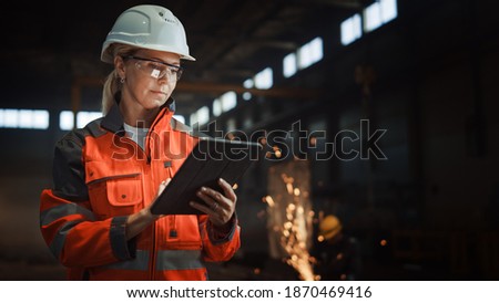 Professional Heavy Industry Engineer Worker Wearing Safety Uniform and Hard Hat Uses Tablet Computer. Serious Successful Female Industrial Specialist Standing in a Metal Manufacture Warehouse. Royalty-Free Stock Photo #1870469416